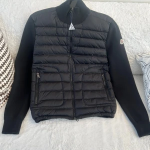 Moncler cardigan svart - Snygg svart cardigan från Moncler med quiltad framsida och stickade ärmar. Jackan har hög krage, två dragkedjefickor och Moncler-logga på ena ärmen. Perfekt för dig som vill ha en stilren och exklusiv look under kyliga dagar. Jackan har aldrig använts