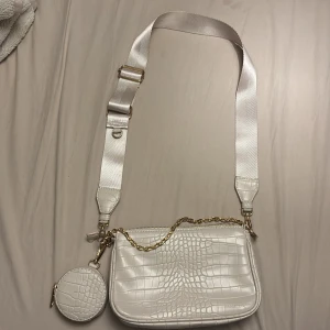 Vit axelväska med krokopräglat mönster - Snygg vit axelväska i krokopräglat syntetmaterial med bred axelrem och guldfärgade detaljer. Väskan har två fack med dragkedja och en kedja framtill samt en matchande rund miniväska som hänger på sidan. Perfekt för att lyfta din outfit.
