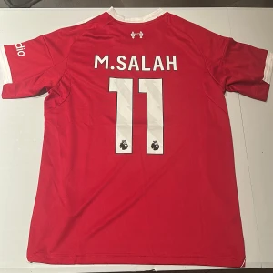 Liverpool M. Salah 11 Adidas matchtröja - Liverpool FC fotbollströja med M. Salah och nummer 11 på ryggen. Röd med vita detaljer, Adidas-logga och klubbmärke på bröstet. Standard Chartered som sponsor framtill, Premier League-märke på ärmen. Tillverkad i lätt och ventilerande material. Replika, endast handtvätt gäller!