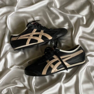 Onitsuka Tiger svarta sneakers - Svarta Onitsuka Tiger sneakers med beigea detaljer och klassiska ränder på sidorna. Skorna har snörning, platt sula och rund tå. Tillverkade i skinn och mocka med vita kanter runt öppningen. Perfekta för dig som gillar retrostil och streetwear.