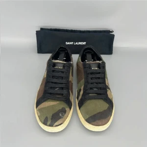 Saint Laurent sneakers i camouflage - Saint Laurent sneakers med camouflage-mönster i grönt, brunt och svart. Skorna har svarta snören, beige sula och Saint Laurent-logga på sidan. Tillverkade i canvas med detaljer i mocka. Perfekta för dig som vill sticka ut med en cool streetstyle.