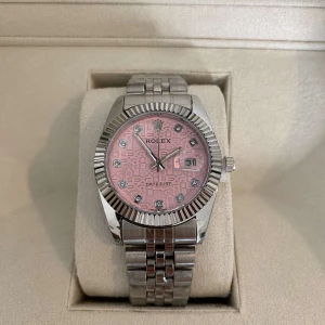 Datejust med rosa urtavla - Säljer en snygg Datejust med silverfärgad länk och boett i stål. Urtavlan är rosa med mönster och glittrande indexmarkeringar. Klockan har räfflad bezel och datumfönster vid kl 3. Kommer i originalbox med certifikat och tillbehör.