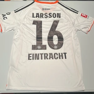 Eintracht Frankfurt Larsson 16 Adidas tröja - Snygg vit Eintracht Frankfurt matchtröja från Adidas med Larsson och nummer 16 på ryggen. Tröjan har svarta detaljer, orange kantband och klubbmärke på bröstet. Tillverkad i lätt och ventilerande material med AEROREADY-teknologi. Perfekt för fotbollsfans! Replika, endast handtvätt gäller!