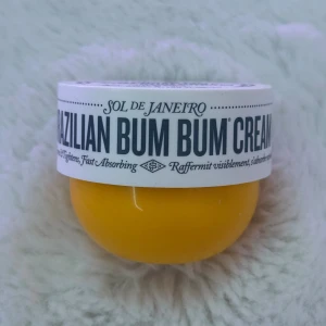 Sol de Janeiro Brazilian Bum Bum Cream - Ikonisk Brazilian Bum Bum Cream från Sol de Janeiro i en rund, gul plastburk med vit skruvlock. Doft av Cheirosa 62, berikad med guaranáextrakt och koffein. Perfekt för dig som älskar lyxiga, väldoftande kroppskrämer med tropisk vibe.