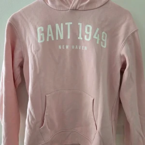 Rosa hoodie från GANT med tryck - Snygg ljusrosa hoodie från GANT med vit text 'GANT 1949 New Haven' på bröstet. Tröjan har huva, magficka och långa ärmar. Mjuk bomullskvalitet som är skön att ha på sig. Perfekt för chill eller häng med kompisar.