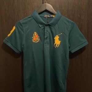 Grön pikétröja från Polo Ralph Lauren - Snygg grön pikétröja från Polo Ralph Lauren med klassisk krage och knappar. Stora gula broderade loggor på bröstet och ärmen ger en sportig vibe. Perfekt för dig som gillar stilrena och ikoniska plagg med premiumkänsla.