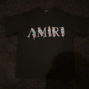 Svart blommig t-shirt från Amiri - Svart t-shirt från Amiri med stort tryck framtill där märkesnamnet är dekorerat med färgglada blommor i blått, rosa och gult. Klassisk passform och korta ärmar. Perfekt statement-plagg för dig som gillar streetwear med en twist.