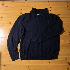 Mörkblå stickad tröja Polo Ralph Lauren - Mörkblå stickad tröja från Polo Ralph Lauren med ribbad hög krage och två knappar vid halsen. Klassisk broderad logga på bröstet. Tröjan är långärmad och har breda ribbade muddar vid ärmslut och nederkant. Tillverkad i mjuk bomull. Extremt sällsynt och 5000 i nypris. L men passar M
