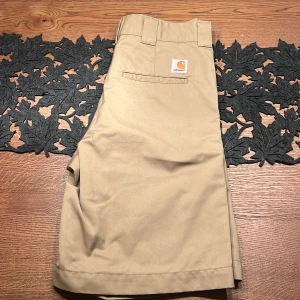 Beige shorts från Carhartt - Säljer ett par klassiska beige chino shorts från Carhartt med raka ben och baggy passform. De är köpta i Tyskland i carhartt butik. De har bälteshällor, dragkedja och fickor både fram och bak. Perfekta för en chill sommarstil och tillverkade i slitstark bomull.