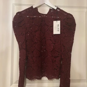 Vinröd spetsblus från NA-KD - Säljer en helt ny med prislapp vinröd långärmad blus från NA-KD i vacker spets med blommigt mönster. Blusen har rund halsringning och transparenta partier som ger en snygg look. 