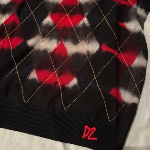 Svart rutig stickad tröja från Dsquared2 - Stickad svart tröja från Dsquared2 med rutigt mönster i rött, vitt och grått samt tunna gula linjer. Snygg broderad röd D2-detalj nertill. Nypris 3600kr, barnstorlek 14Y men passar vuxen XS
