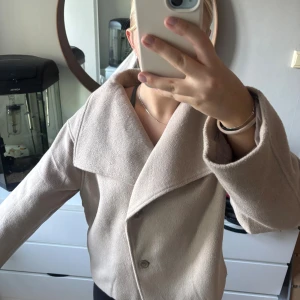 Beige kappa med bred krage - Säljer en stilren beige kappa med bred krage och två knappar framtill. Jackan har en rak och lite loose passform, perfekt för dig som gillar en clean och enkel look. Materialet känns mjukt och ger en lyxig känsla. Passar perfekt till höst och vår.
