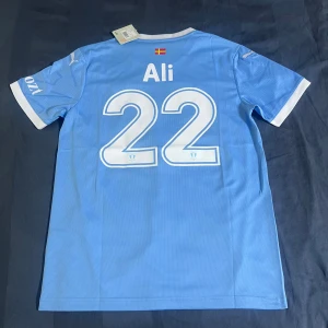 Malmö FF fotbollströja Puma M - Snygg ljusblå Malmö FF fotbollströja från Puma med vita detaljer på krage och ärmslut. Tryck med 'Ali' och nummer 22 på ryggen samt klubbmärke och texten 'Framåt Malmö!' på bröstet. Tillverkad i lätt och ventilerande polyester med RE:FIBRE-teknologi.