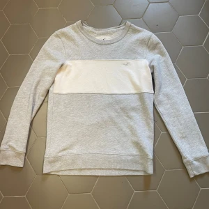 Grå och vit tröja från Hollister  - Snygg grå tröja från Hollister i storlek XS med vit panel över bröstet och diskret broderad logga. Tröjan har rund halsringning och långa ärmar. Perfekt för en chill och stilren look.