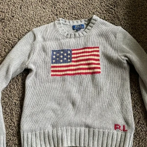 Grå stickad tröja Polo Ralph Lauren - Grå stickad tröja från Polo Ralph Lauren med amerikansk flagga på bröstet och RL-broderi nertill. Klassisk rund halsringning och ribbade muddar vid ärmslut och nederkant. Perfekt för dig som gillar preppy stil.