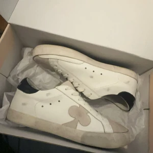 The Gallerist skor  - Säljer ett par vita sneakers med beige mockadetalj i form av en klöver på sidan. Skorna har snörning, rund tå och svart hälparti. Snygg och stilren design som passar till det mesta. Tillverkade i skinn och mocka för en schysst look.
