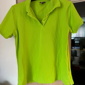 Limegrön pikétröja från ID game - Fräsch limegrön pikétröja från ID game  i storlek L. Klassisk krage, korta ärmar och två knappar framtill. Tillverkad i mjuk bomull som känns skön mot huden. Perfekt för dig som gillar färgstarka plagg och vill sticka ut lite extra.