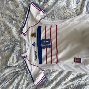 Frankrike Zidane 10 VM 98 Adidas tröja - Klassisk vit fotbollströja från Adidas, Frankrikes landslag VM 1998 med Zidane 10 på ryggen. Tröjan har blå och röda detaljer, tunna röda ränder framtill och broderade märken. Kortärmad modell i lätt och ventilerande material.