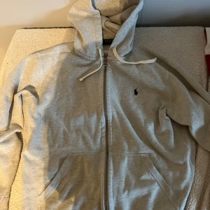 Grå hoodie från Polo Ralph Lauren - Snygg grå hoodie från Polo Ralph Lauren med klassisk broderad logga på bröstet. Tröjan har dragkedja, huva med snörning och två fickor framtill. Tillverkad i mjuk bomull, perfekt för chill dagar. Passformen är normal och ärmarna är långärmade.