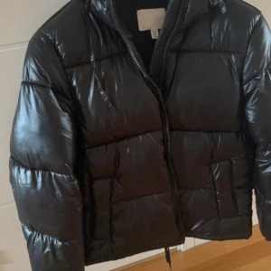 Svart pufferjacka med dragkedja - Säljer en svart pufferjacka med glansig yta och vadderade sektioner. Jackan har hög krage, lång ärm och praktisk dragkedja framtill. Perfekt för kalla dagar och har en modern, oversized look.