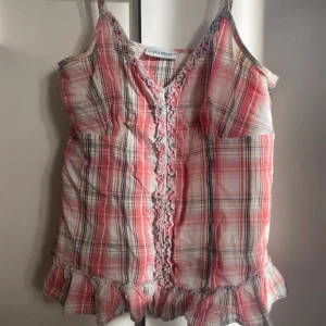 Rutig linnetopp från Clockhouse - Supersöt rutig linnetopp från Clockhouse i rosa, vit och svarta toner. Toppen har smala axelband, volangkant nertill och volangdetaljer längs v-ringningen. Perfekt för varma dagar och ger en lätt och luftig känsla.