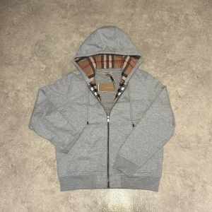 Grå kofta från Burberry  - Säljer en grå hoodie från Burberry i 100% bomull. Klassisk dragkedja framtill, huva med snörning och ikoniskt rutigt foder i huvan. Perfekt för dig som vill ha en stilren och lyxig vardagslook. Storlek M.