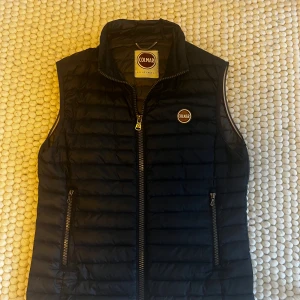 Dunväst från Colmar - Svart quiltad dunväst från Colmar med hög krage och dragkedja framtill. Västen har två fickor med dragkedja och Colmar-logga på bröstet. Perfekt för lager-på-lager och snygg till både hoodie och t-shirt.