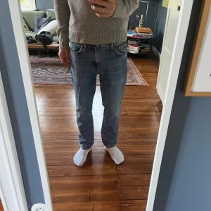 Blå jeans från Nudie med raka ben - Säljer ett par klassiska blå jeans från Nudie med raka ben och normal passform. Jeansen har fem fickor, snygga kontrastsömmar och Nudie-logga på bakfickan. Tillverkade i slitstark denim av bomull. Perfekta för dig som gillar en enkel och tidlös look. MODELL: gritty Jackson 