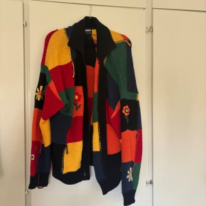 Färgglad cardigan jacka från Minga - Säljer en stickad cardigan jacka från Minga med patchwork-design i rött, gult, grönt, blått och orange. Jackan har blommiga broderier, lång ärm och en bred krage. Perfekt för dig som gillar färg och unika detaljer!