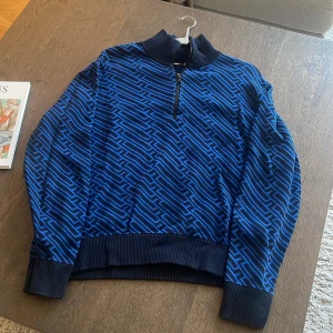 Blå mönstrad half zip - J.Lindeberg - Blå och svart mönstrad tröja från J.Lindeberg med ribbad krage och muddar. Tröjan har en half zip-dragkedja och ett grafiskt mönster över hela plagget. 