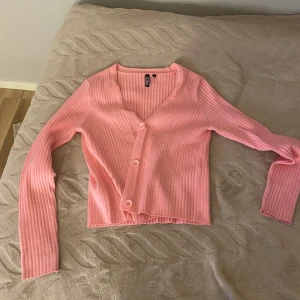 Rosa ribbad kofta från Pieces XS - Superfin rosa kofta från Pieces i storlek XS. Koftan är ribbstickad med v-ringning och tre knappar framtill. Långärmad och croppad modell som ger en trendig look. Perfekt att slänga över en topp för en söt och avslappnad stil.