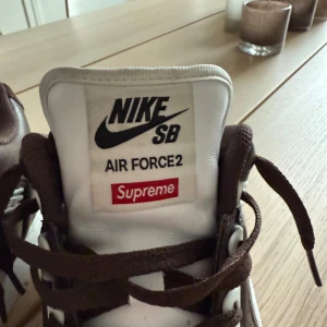 Nike SB Air Force 2 x Supreme bruna - Nike SB Air Force 2 Supreme i brunt och vitt skinn med klassisk Supreme branding på sidan och röd innersula. Låga sneakers med snörning, vit sula och perforerade detaljer. Perfekta för dig som gillar streetwear och unika collabs. Storlek 45,5