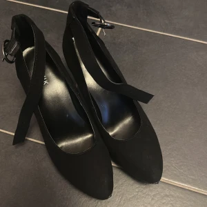 Svarta pumps med platå från Catwalk - Snygga svarta pumps från Catwalk med spetsig tå och platåsula. Skorna har en bred rem med spänne runt vristen och är gjorda i mockaliknande material. Perfekta för dig som vill ha en stilren och trendig look med lite extra höjd.