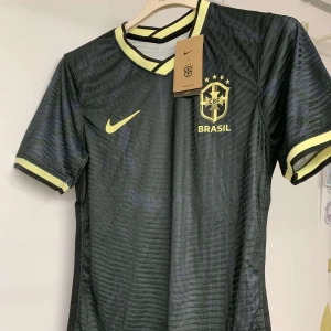 Svart Brasilien fotbollströja Nike - Snygg svart Brasilien fotbollströja från Nike med gula detaljer och CBF-emblem på bröstet. Tröjan har korta ärmar, rund hals med gul kant och är tillverkad i ett lätt, ventilerande material som passar perfekt till träning eller match.