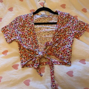 Blommig croppad omlottopp med knyt - Superfin croppad topp med blommigt mönster i rosa, rött och grönt. Toppen har korta ärmar, v-ringning och knyt framtill för en söt och somrig look. Perfekt för varma dagar och lätta outfits.