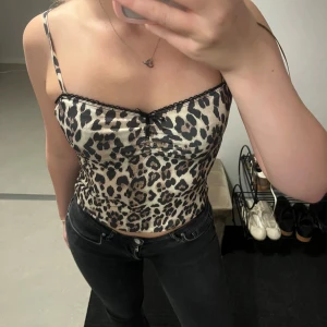 Leopardmönstrad linne-topp med spets - Säljer en snygg leopardmönstrad linne-topp med tunna axelband och liten svart rosett framtill. Toppen har svarta spetsdetaljer längs kanten och är figurnära med croppad passform. Perfekt för dig som vill sticka ut med ett djurmönster!