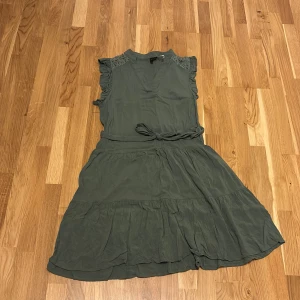 Grön kort klänning  - Supersöt militärgrön kortklänning från Vero Moda i storlek XS. Klänningen har volangdetaljer vid axlarna, knytband i midjan och är ärmlös. Den har en lätt v-ringning och är tillverkad i ett mjukt, tunt material som känns luftigt och skönt. Bara använd en gång💕