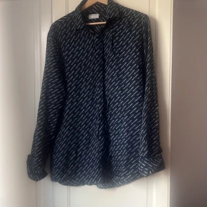 Mörkblå skjorta Tiger of Sweden - Snygg mörkblå skjorta från Tiger of Sweden med vit logotyp-print över hela tyget. Slim fit-modell med långa ärmar och klassisk krage. Skjortan har knäppning framtill och är tillverkad i ett mjukt, lätt material som känns skönt mot huden.