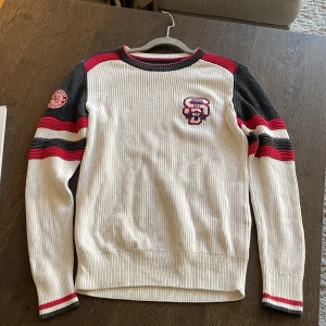 Vit och röd stickad tröja - Superdry - Stickad tröja från Superdry i vintage-stil, vit med mörkgrå och röda detaljer på ärmar och hals. Broderad Superdry-logga på bröstet och patch på ena ärmen. Ribbad struktur och rund hals. 