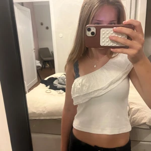 Halfshoulder topp med volang - Säljer en vit ribbad topp med oneshoulder-design och bred volang med broderade blommor. Toppen är ärmlös på ena sidan och har en snygg asymmetrisk form. Perfekt för dig som gillar unika detaljer och somrig stil.