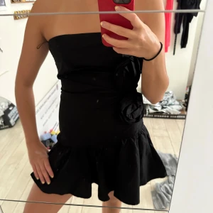 As snygg klänning från zara - Supersnygg svart axelbandslös kortklänning med volang nertill och två stora tygrosor på sidan. Klänningen har en figurnära passform och är perfekt för dig som vill sticka ut. Materialet känns mjukt och följsamt mot kroppen. Prislappen är fortfarande på!