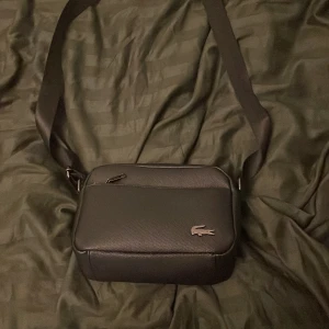 Svart axelväska från Lacoste - Snygg svart axelväska från Lacoste med klassisk krokodil-logga i metall på framsidan. Väskan har en justerbar axelrem, dragkedja upptill och ett extra fack med dragkedja på baksidan. Tillverkad i slätt syntetmaterial med stilren och enkel design.