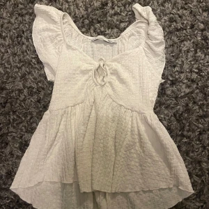 Vit volangblus med knyt från 157 - Supersöt vit blus från 157 i storlek XS. Den har volangiga korta ärmar, knytning framtill och en lätt rynkad struktur i tyget. Blusen har en luftig och flowy passform med extra volang nertill, perfekt för en fräsch och somrig look.