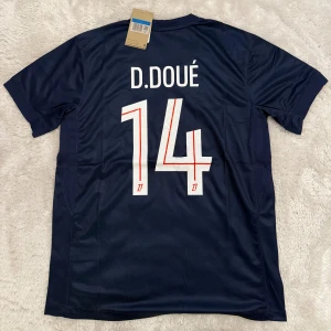 PSG matchtröja D. Doué nr 14 Nike - Paris Saint-Germain fotbollströja med D. Doué och nummer 14 på ryggen. Mörkblå med röd och vit detalj på framsidan, PSG-emblem och Nike-logga. Tillverkad i lätt och ventilerande material, perfekt för fotbollsplanen.