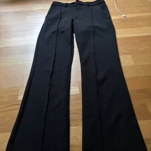 Svarta vida kostymbyxor och normal hög midja - Snygga svarta kostymbyxor med vida ben och pressveck framtill. Byxorna har hällor för bälte och klassisk passform med rak midja. Perfekta för en clean och stilren look. Materialet känns mjukt och följsamt, troligtvis polyesterblandning.