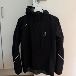 Svart vindjacka från Haglöfs med huva - Svart vindjacka från Haglöfs med huva och reflekterande detaljer på ärmarna. Jackan har en minimalistisk design med diskret logga på bröstet och dragkedja framtill. .
