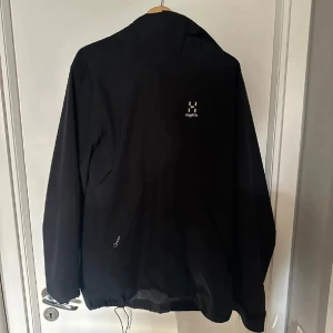Svart vindjacka från Haglöfs med huva - Svart vindjacka från Haglöfs med minimalistisk design och praktisk huva. Jackan har diskret logga på bröstet och justerbar nederkant med dragsko. Perfekt för blåsiga dagar och outdoor-aktiviteter. Lätt och smidig att bära med sig.