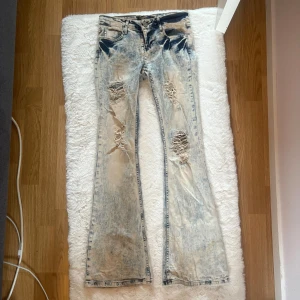 y2k acid wash bootcut jeans med slitningar - Säljer ett par lowrise vintage y2k ljusa acid wash jeans från med bootcut passform. Byxorna har flera slitningar och hål på framsidan för en riktigt edgy look. Perfekta för dig som gillar en retro vibe och vill sticka ut.