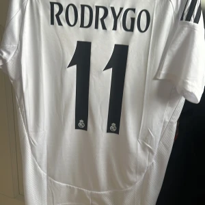 Real Madrid Rodrygo 11 Adidas tröja - Säljer en  Real Madrid matchtröja från Adidas med Rodrygo och nummer 11 på ryggen. Tröjan är vit med svarta detaljer, klubbmärke på bröstet och tre svarta ränder på axlarna. Materialet är lätt och andas, perfekt för fotboll eller supporterliv.