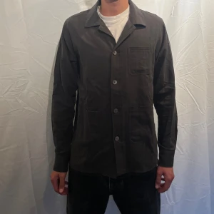  overshirt från TS Studios - Olivgrön overshirt från TS Studios i storlek S. Skjortan har klassisk krage, tre framfickor och knäppning med fyra knappar. Materialet är mjukt och känns som bomull, perfekt till höst för lager-på-lager. Enkel och stilren design som funkar till allt